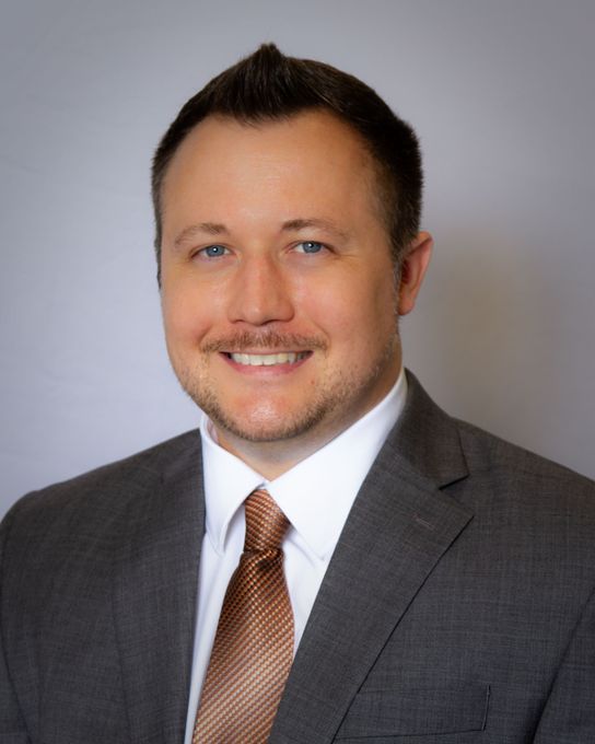 Man in gray suit, white shirt, and brown tie smiling, looking at the viewer.