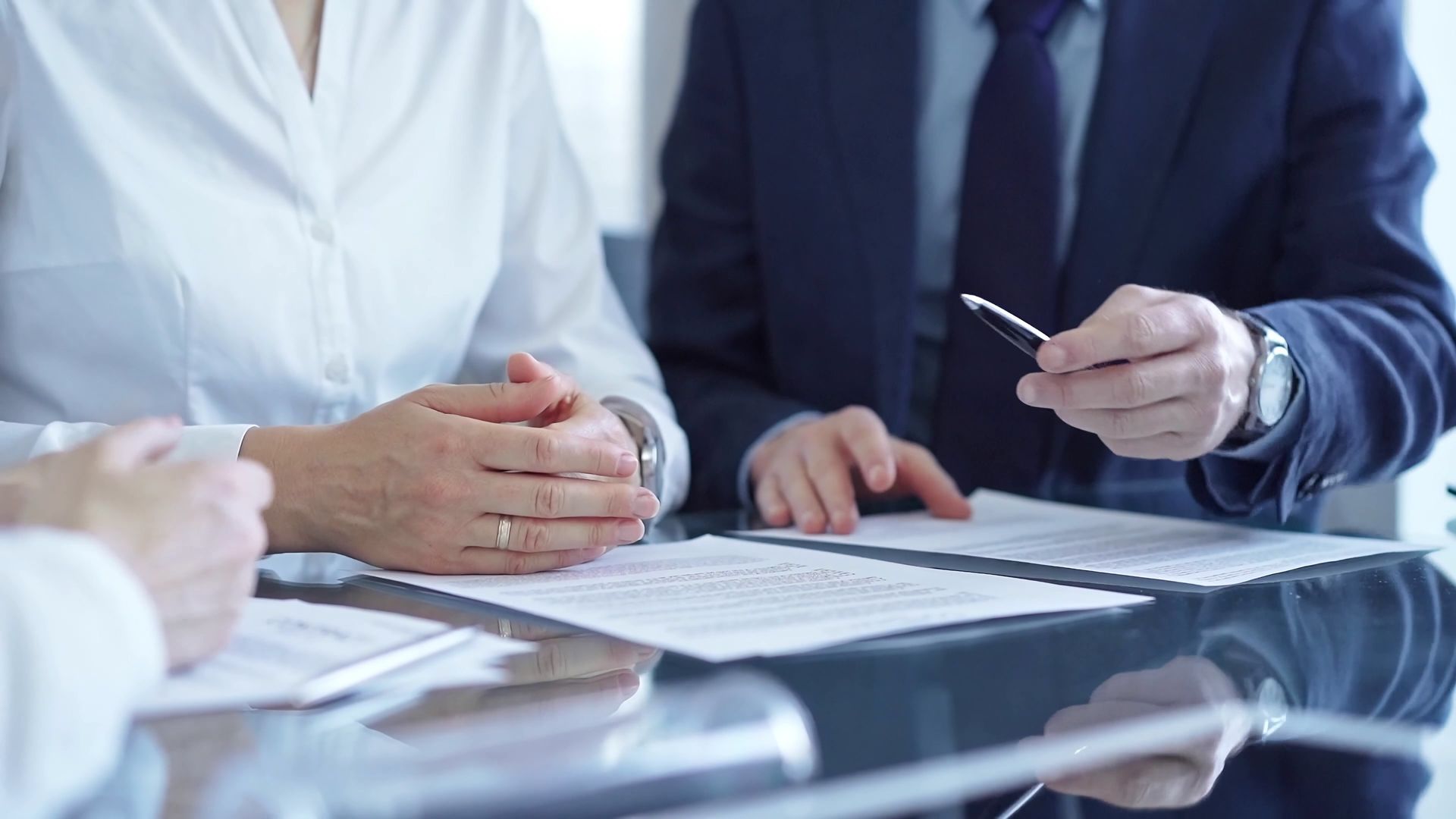 Businesspeople review documents at a table. A man in a suit points a pen at a paper.