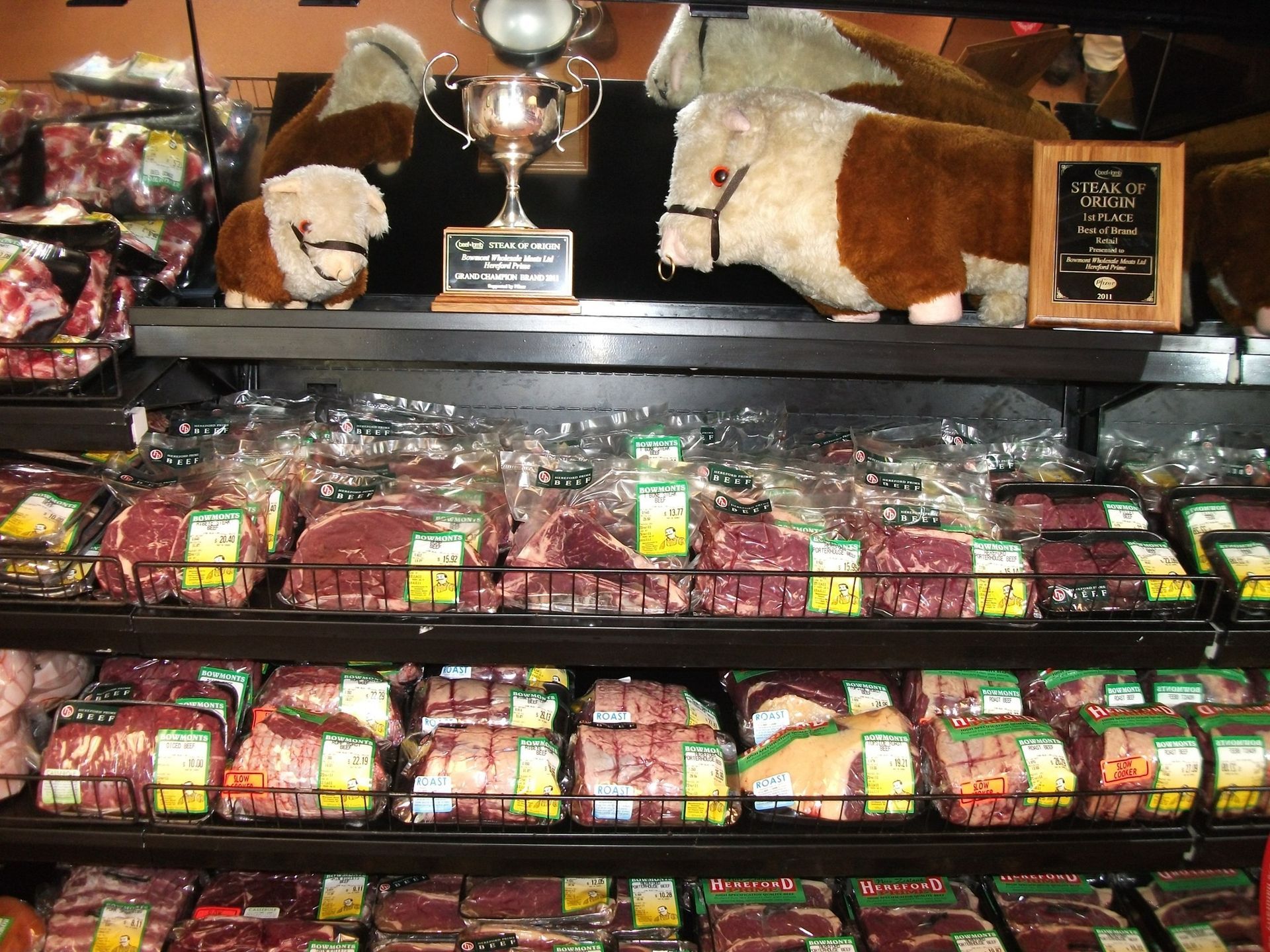 Meat display at a grocery store, with stuffed cows, trophy and signage on the top shelf.