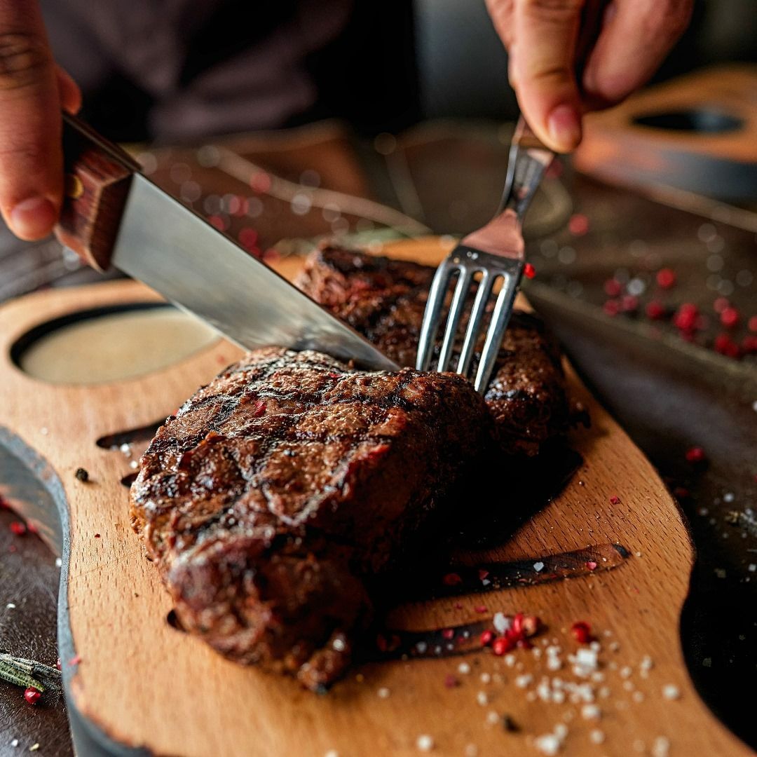 Person cutting steak with knife and fork on a wooden cutting board.