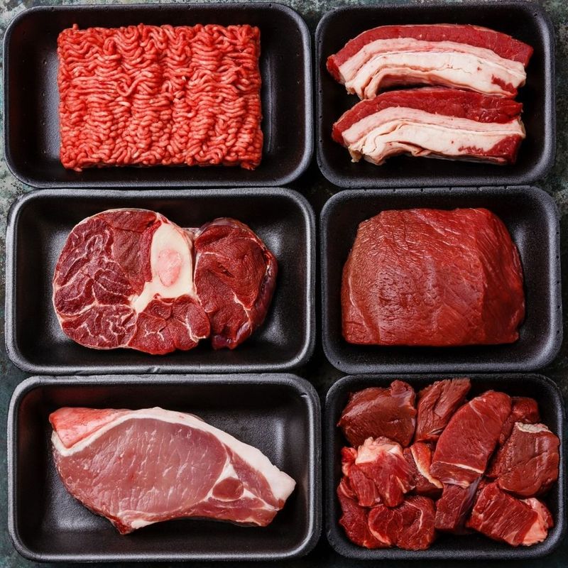 Various cuts of raw meat in black trays: ground beef, bacon, bone-in steak, steak, pork chop, diced meat.