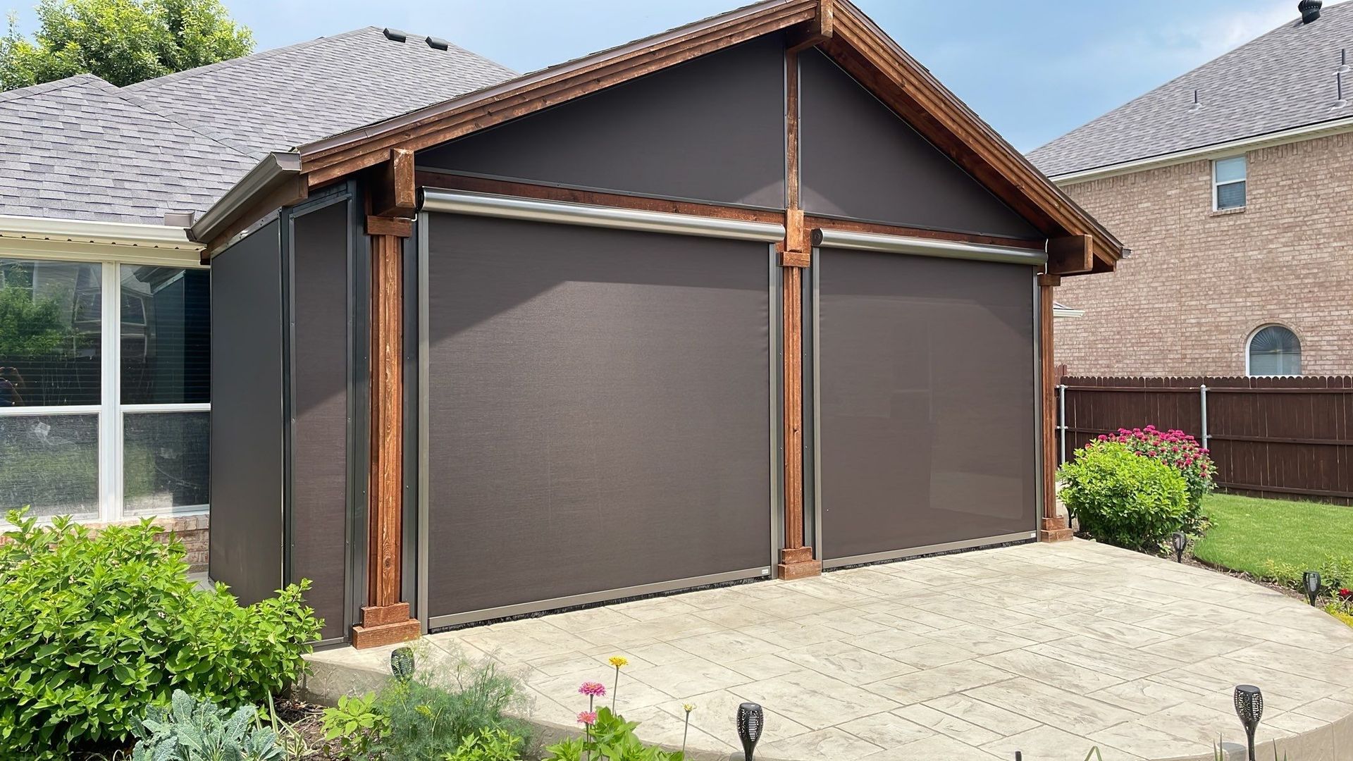 Brown screened-in patio with wooden trim and roller shades, on a brick patio, next to a house with a tile roof.