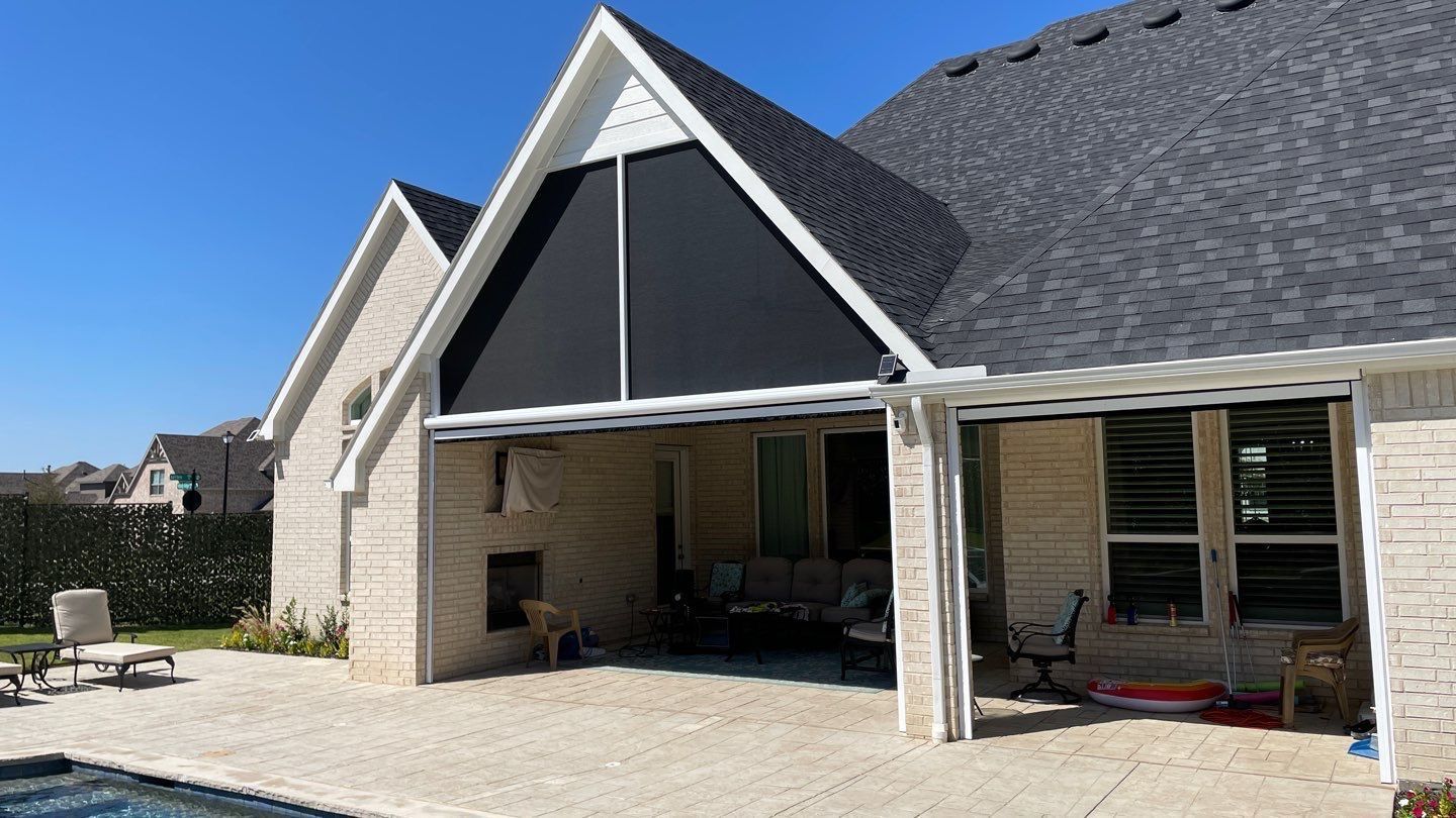 Backyard patio with retractable black sun shade, tan brick house, blue sky.
