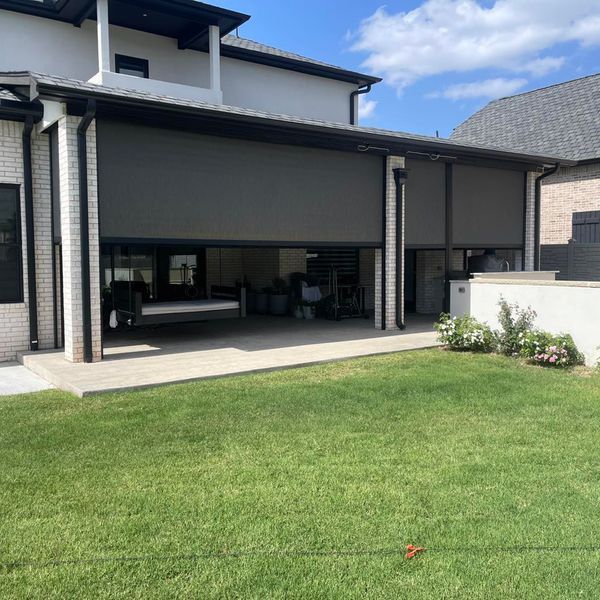 Outdoor patio with retractable dark gray shades, overlooking a grassy lawn. The house has white brick and a black roof.