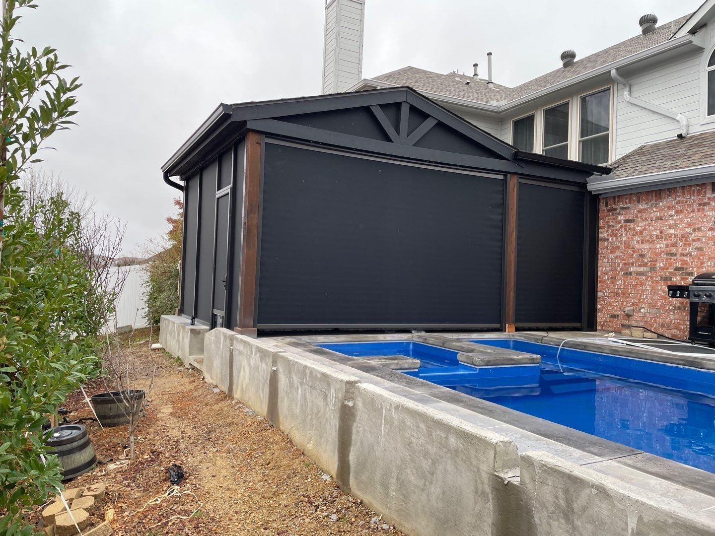 Black-walled outdoor structure with a pool; brick house in the background. Cloudy day.