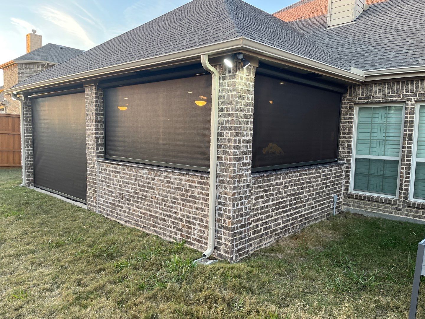 Exterior of brick patio with retractable shades. Brown brick walls, dark shades, green lawn.