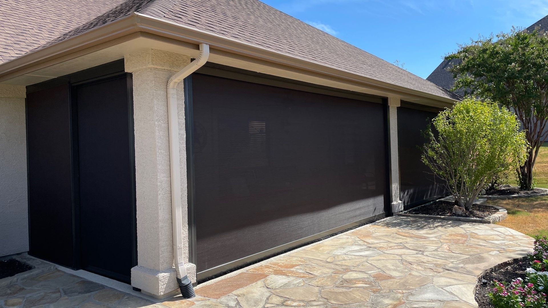 Dark brown outdoor roller shades on a building exterior, providing shade. The building is light-colored with a stone patio in the foreground.