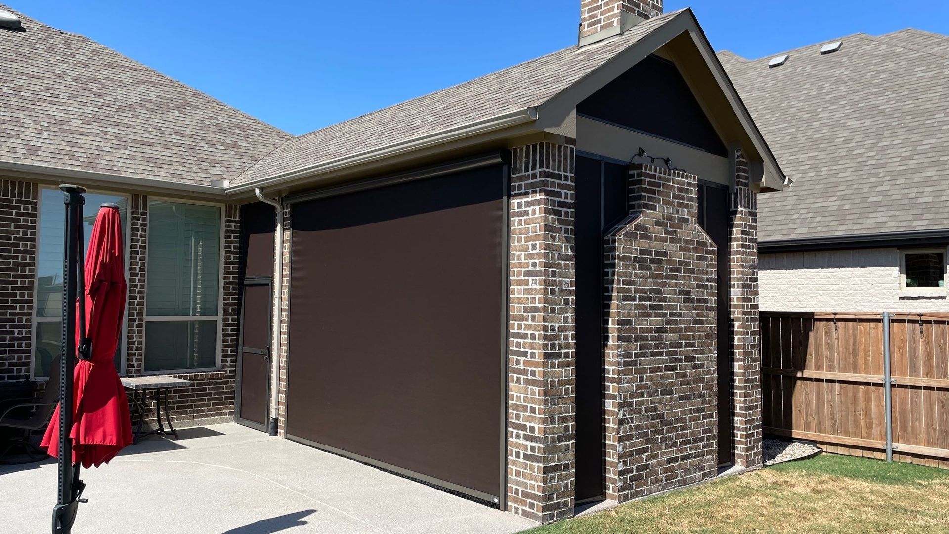 Brown outdoor shade covers an enclosed patio, brick and brown siding, next to a brick chimney and brown roof.
