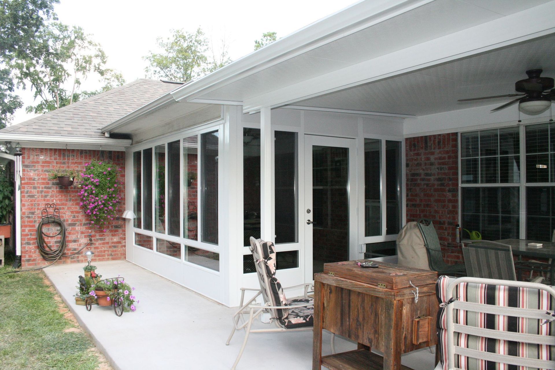 Screened-in porch attached to a brick house with concrete patio, furniture, and a cooler.