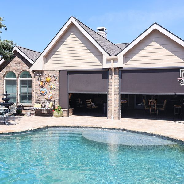 Backyard with a pool, house, and open garage doors with shades. Blue pool, brick and tan house, sunny day.