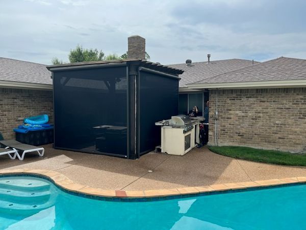 A black screened outdoor kitchen and patio area next to a pool. There is a grill, chimney, and chaise lounge.