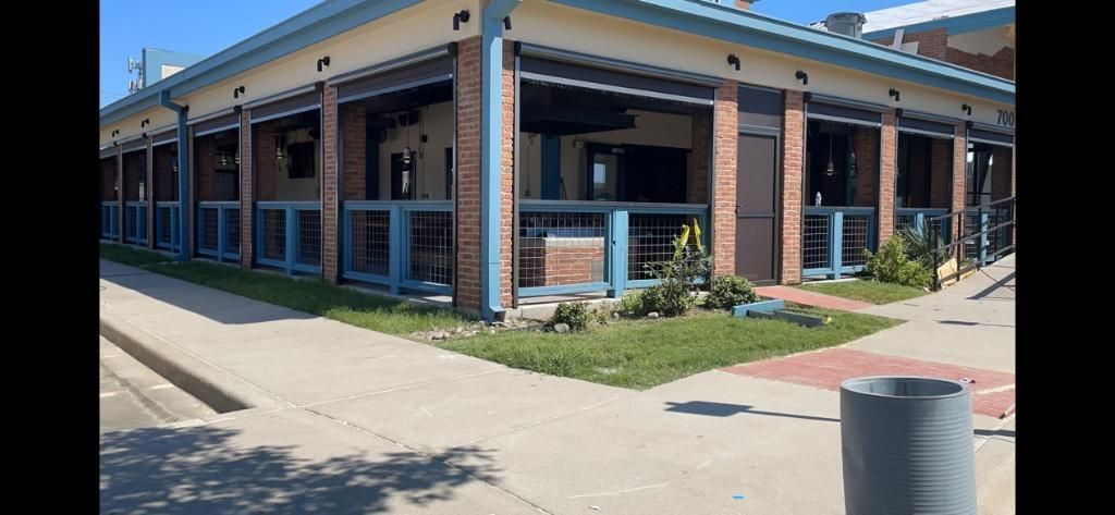 Exterior view of a building with a blue facade, brick pillars, and sidewalk on a sunny day.