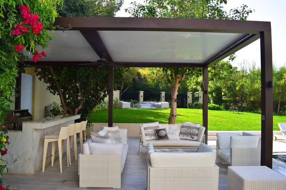 Outdoor patio with white furniture under a brown pergola, bar with stools, green lawn in background.