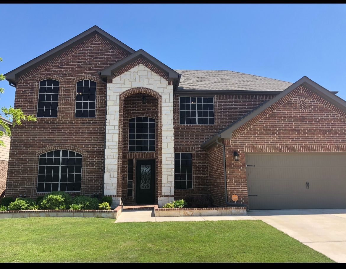 Two-story brick house with a stone entryway, green lawn, and two-car garage under a blue sky.