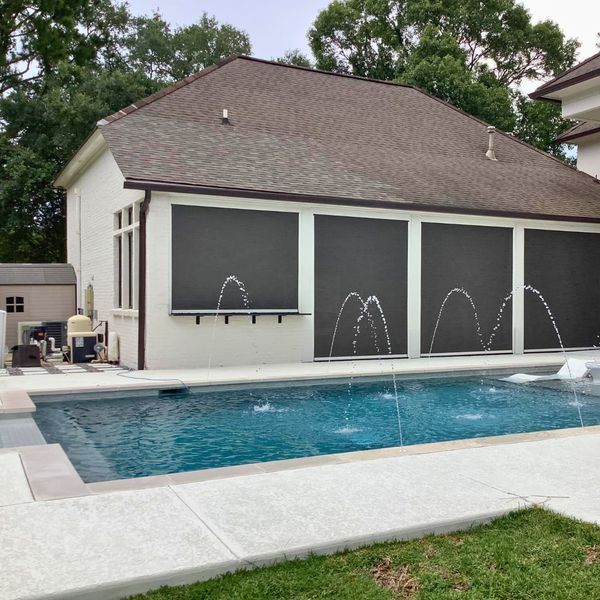 A pool area with a white building featuring dark outdoor screens, a brown roof, and fountains spraying into the water.