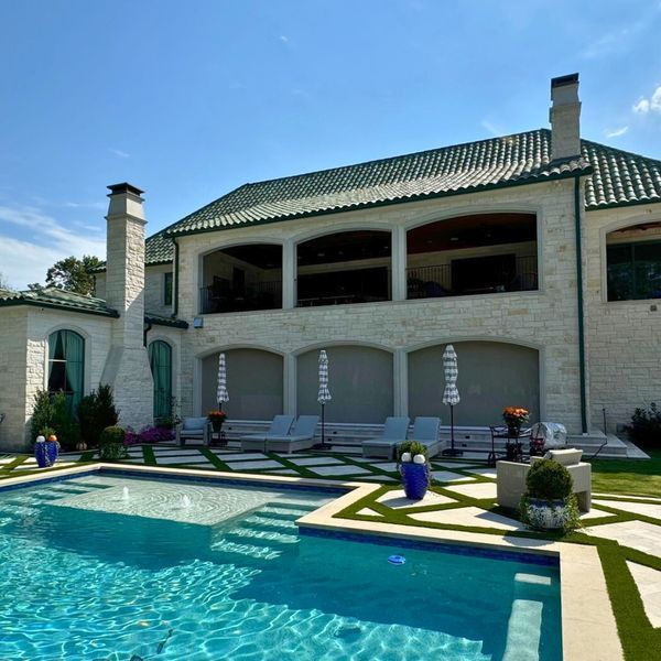 Luxury home with a pool; the house has a green tile roof, a covered patio, and cream-colored stone exterior, blue pool water, and sun loungers.