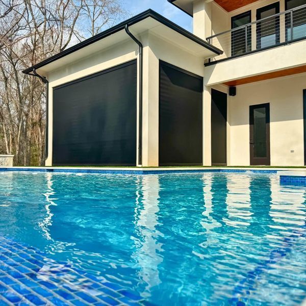 A swimming pool in the foreground with a modern white building with black roller shutters and a balcony in the background.