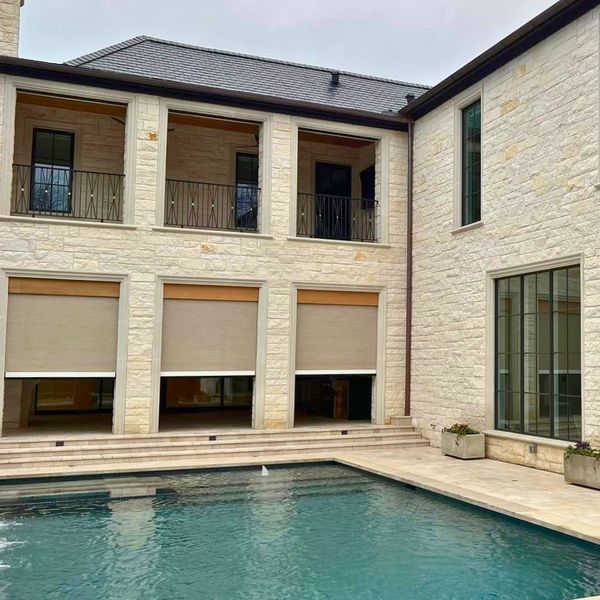 Back of a stone house with a pool. Beige awnings cover ground-floor openings, and a balcony is on the second floor.