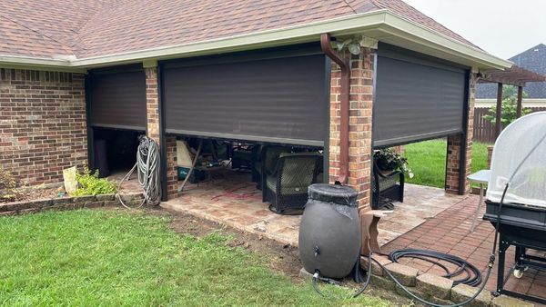 A brown brick patio with partially-closed roll-down shutters, a rain barrel, and outdoor furniture on a rainy day.