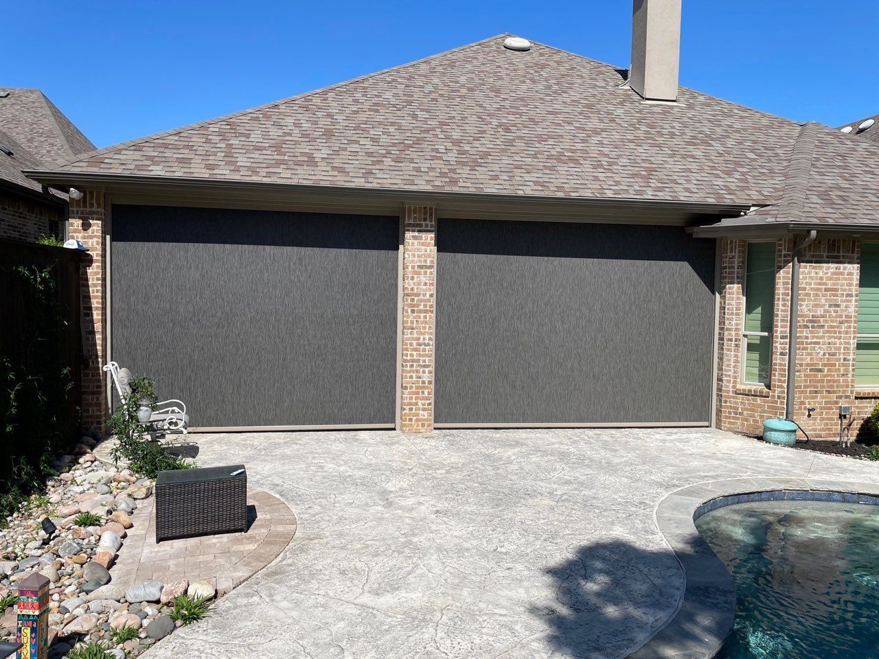 Two dark gray retractable screens cover garage doors of a brick house. A patio with a pool is in the foreground.