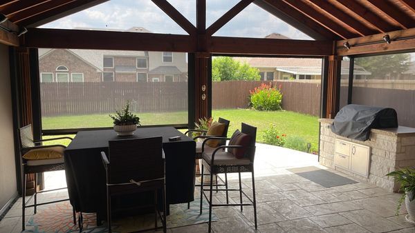 An outdoor screened-in patio with a dining table and chairs, a built-in grill, and a view of a backyard with a wooden fence.