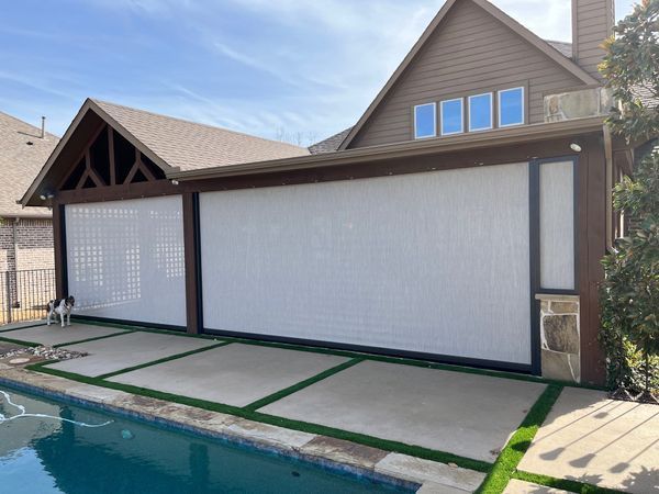 Covered patio with large retractable screens, furniture, and a view of a pool and trees. Black screens are down.