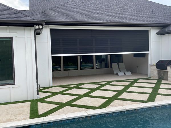 A modern home exterior with a retractable black screen shading an outdoor living space, featuring a pool, turf, and an outdoor grill.