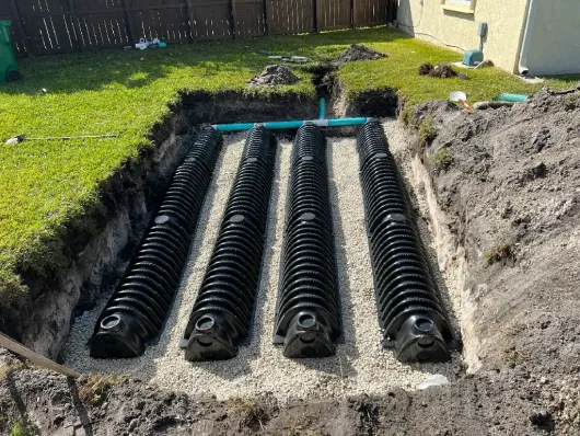 Three black corrugated drainage pipes laid in a trench beside a house, with soil and grass around them