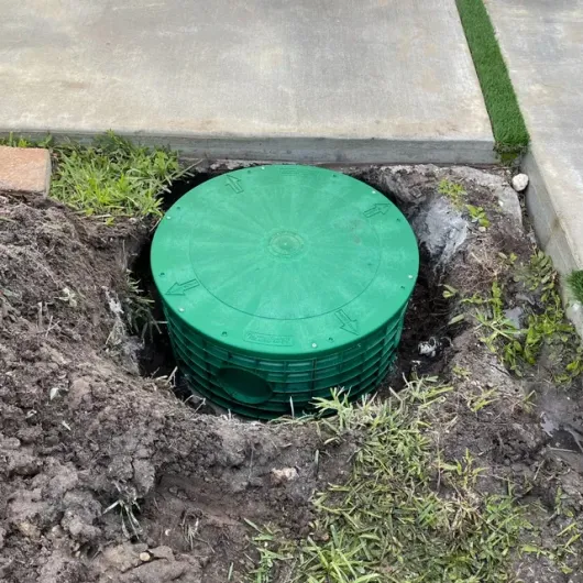 Green utility access cover in a dirt hole beside a concrete sidewalk and grass