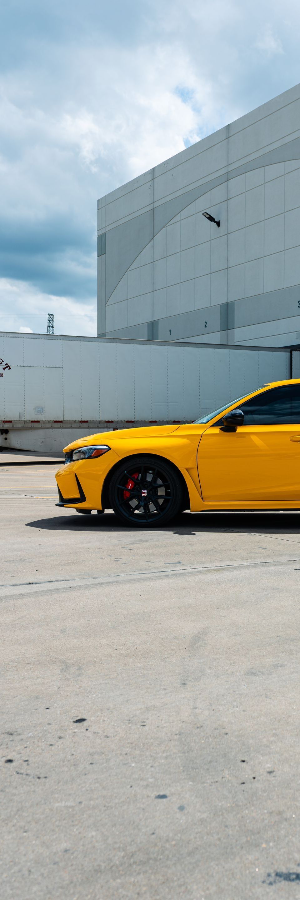 A bright yellow car parked on a concrete surface in front of a large white building against a cloudy sky.