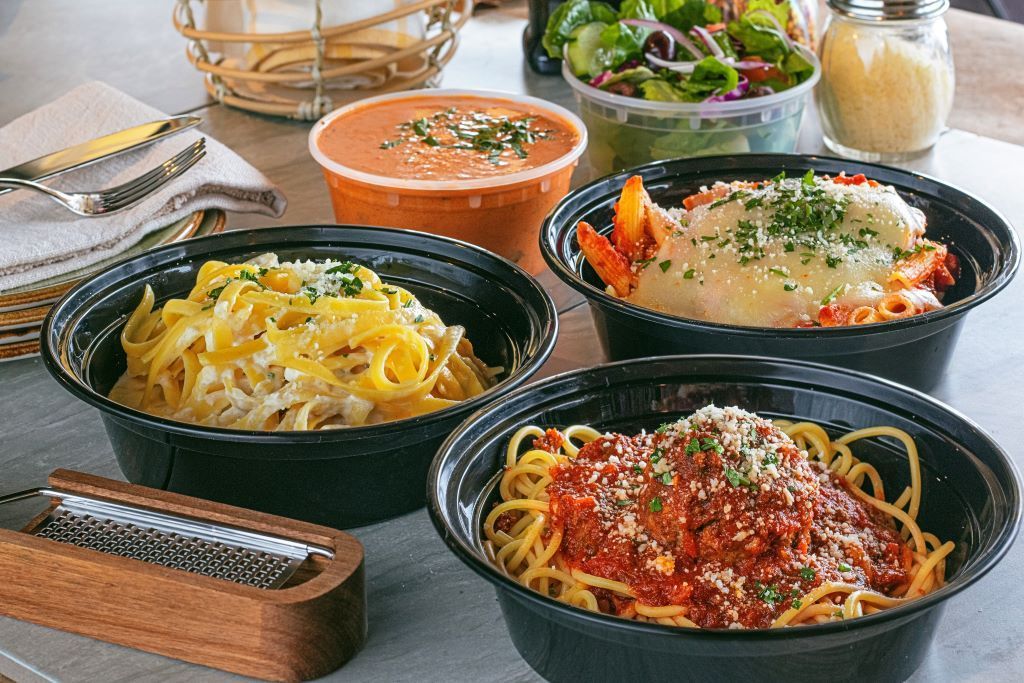 Various pasta dishes in black bowls, plus soup and salad, on a table.