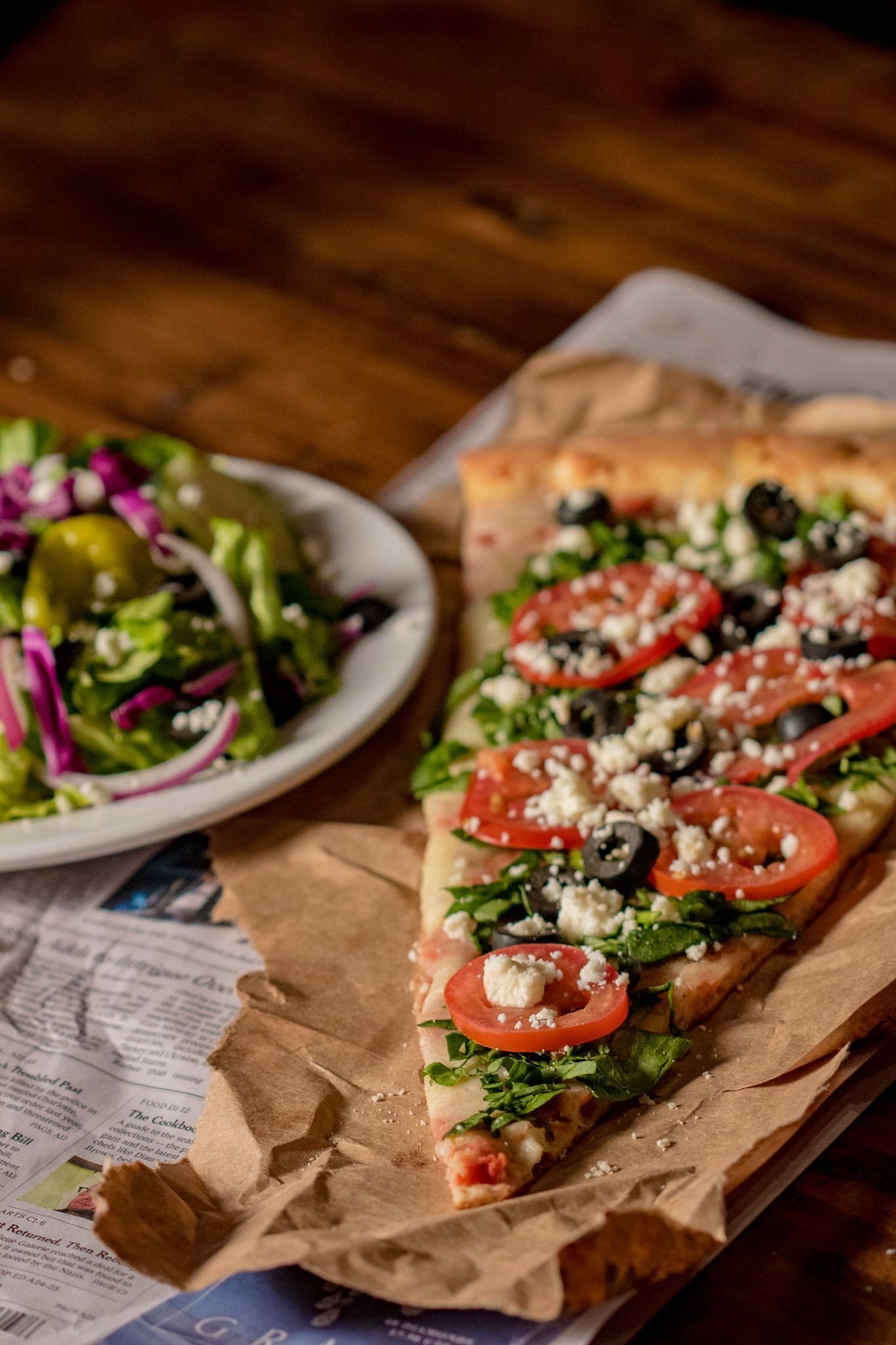 Slice of pizza topped with spinach, tomatoes, olives, and cheese, beside a salad on a white plate, resting on a wood table.