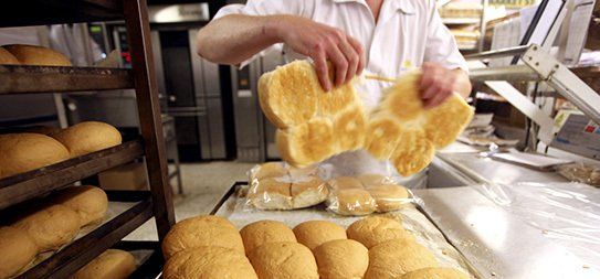 A baker in a white uniform holding up pieces of fresh bread in a bakery setting. Trays of baked goods are visible.