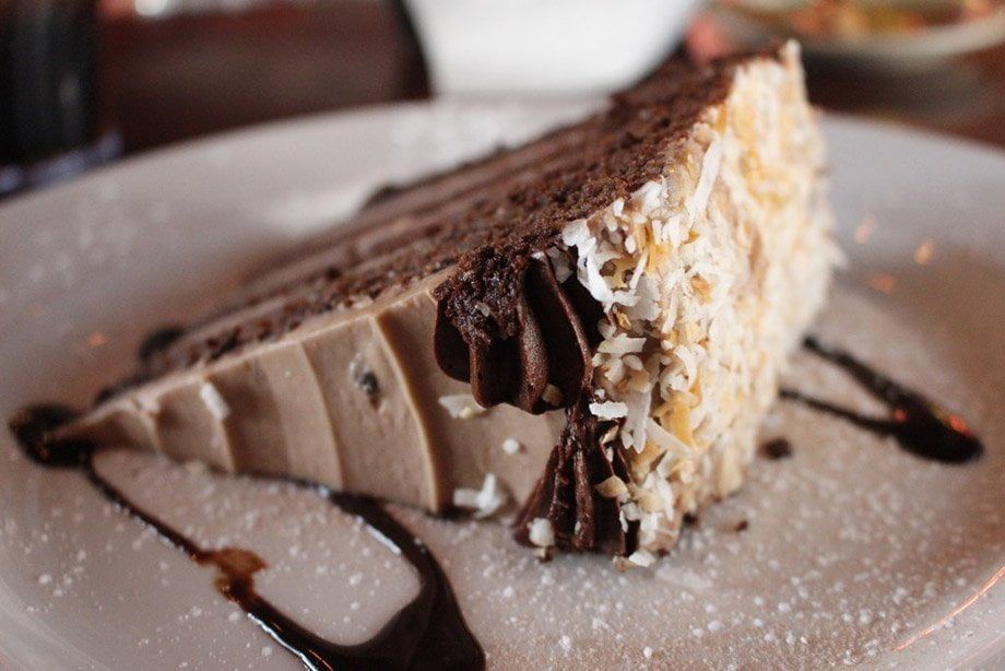 Slice of layered chocolate cake with frosting, coconut, and chocolate drizzle on a white plate.