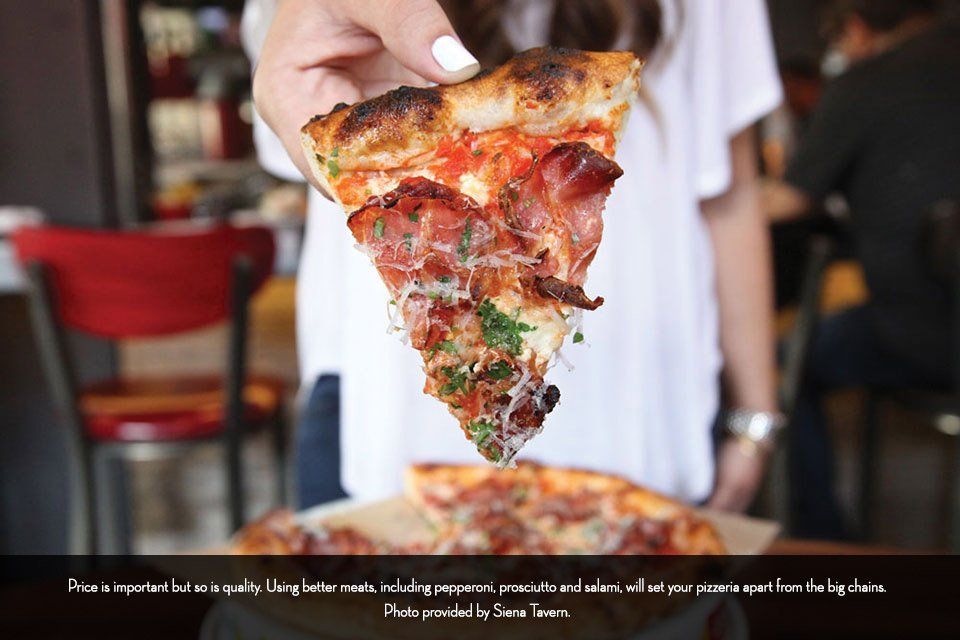 A person holding a slice of pizza with prosciutto and cheese; indoor restaurant setting.