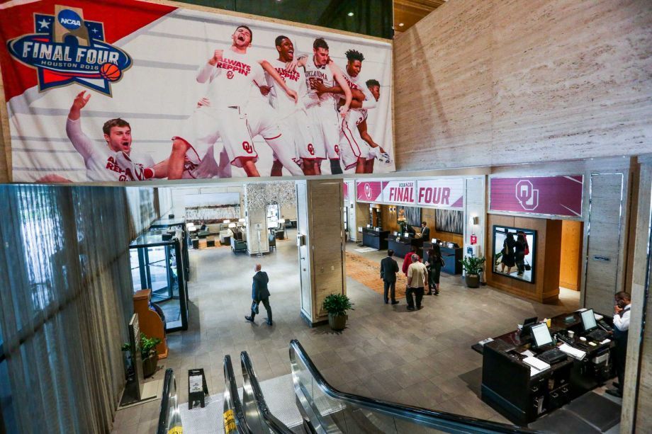 Lobby with a large Final Four banner featuring a basketball team. Several people stand near the entrance and check-in desks. An escalator is in the foreground.