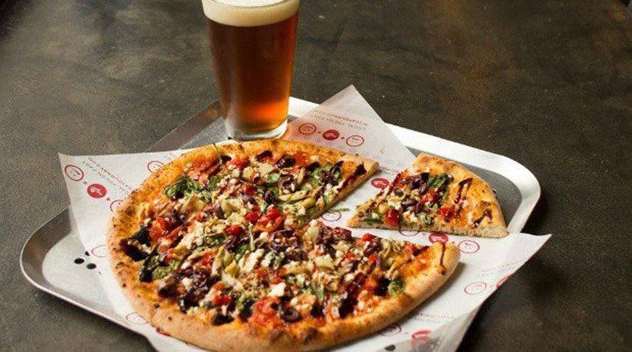 Pizza with a slice removed, on a paper-lined tray, beside a pint of beer. Dark wood table background.