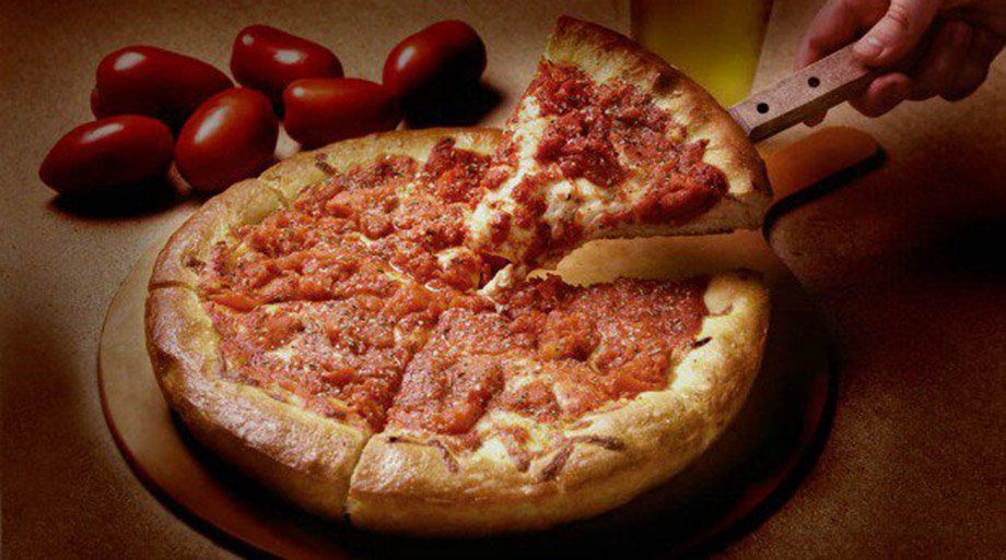 Deep-dish pizza with a slice being lifted by a spatula. Red sauce covers the cheese, and tomatoes are nearby.