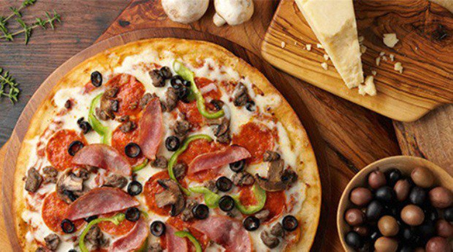 Close-up of a pizza with pepperoni, ham, peppers, olives, and mushrooms on a wooden board, with cheese and olives nearby.