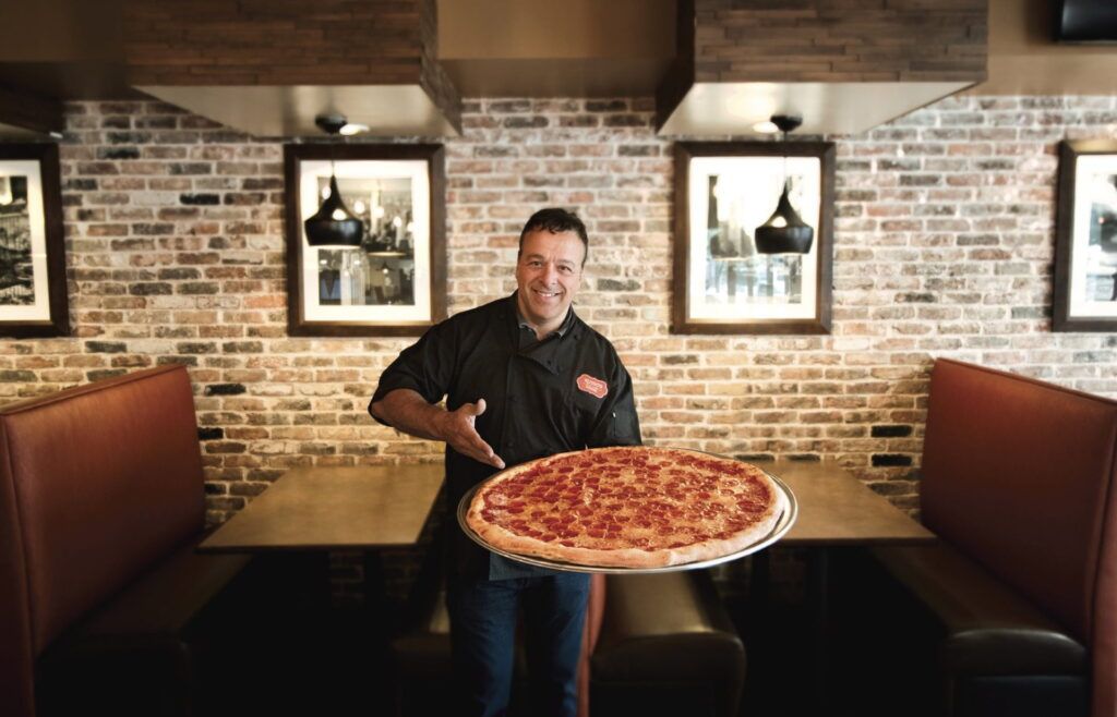 Chef holding a large pizza in a restaurant. He smiles and points at the pizza in a booth-lined area with brick walls and framed photos.