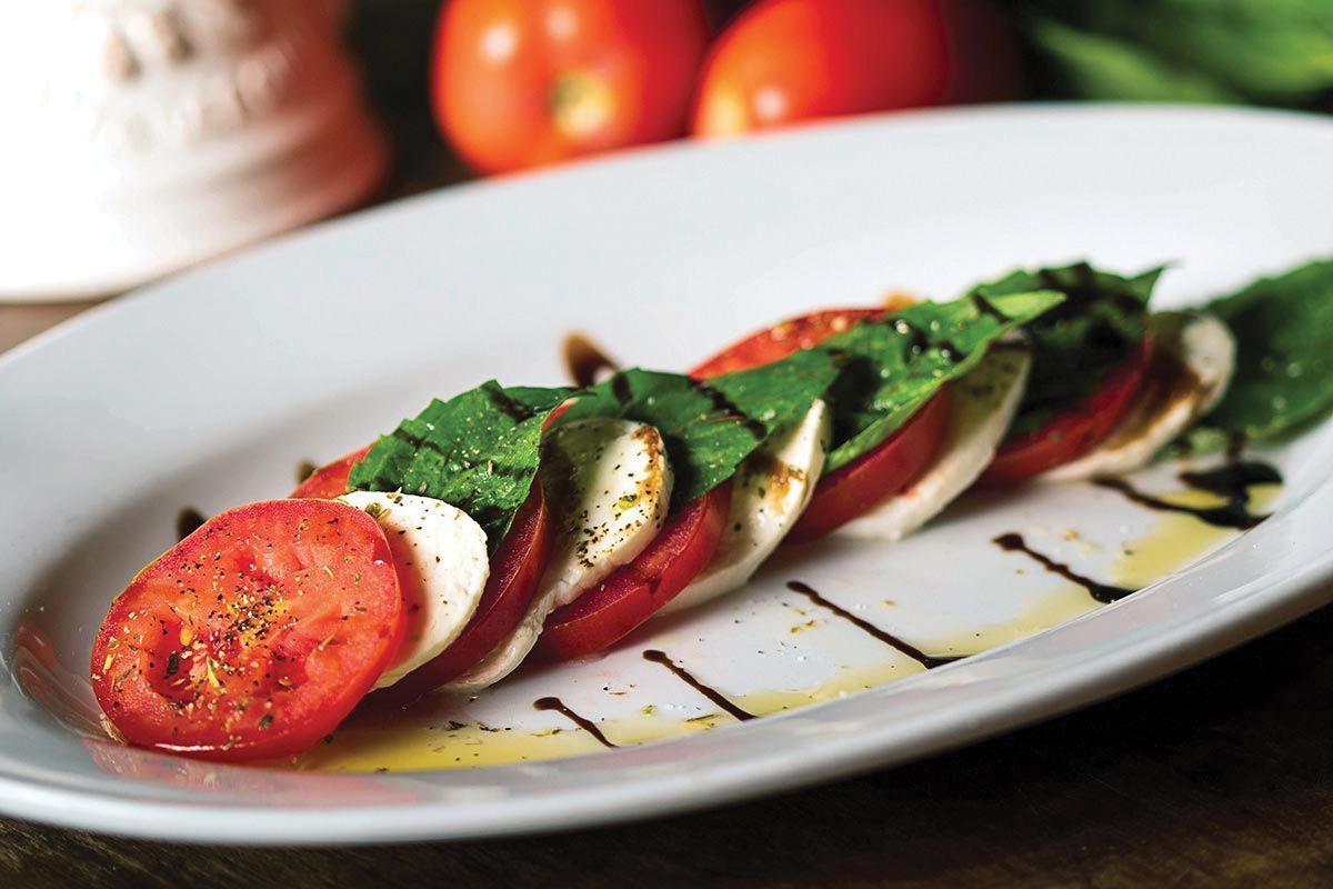 Caprese salad: sliced tomatoes, mozzarella, and basil on a white platter, drizzled with oil and balsamic glaze.