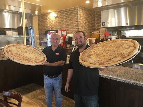 Two men in a pizzeria holding large pizzas. One man smiles, the other has a neutral expression. Behind them is a kitchen.