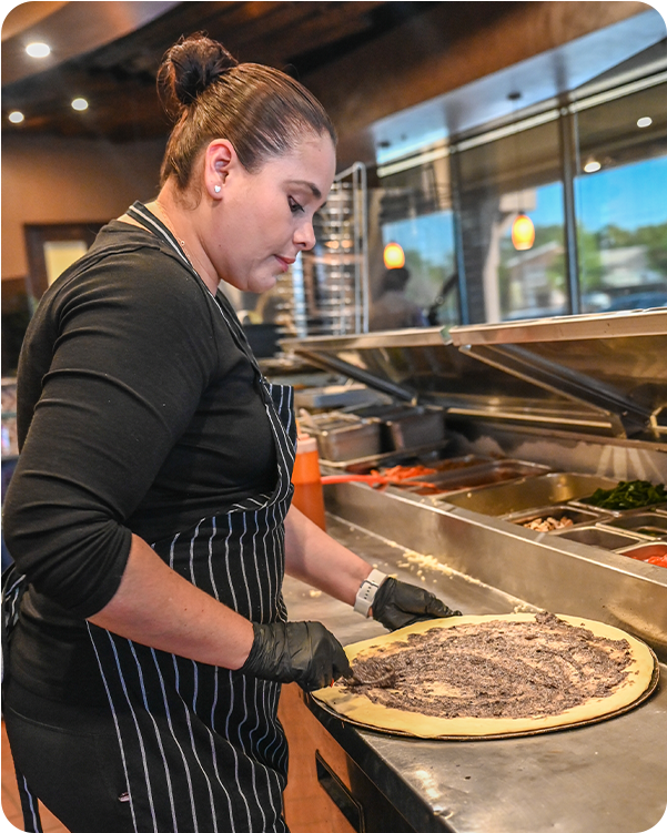Person in black apron spreads topping on pizza in a restaurant kitchen.