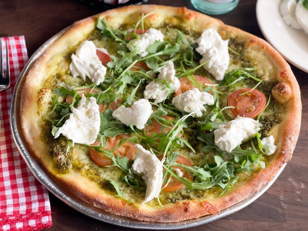 Pizza with pesto base, sliced tomatoes, arugula, and dollops of white cheese on a metal pan.