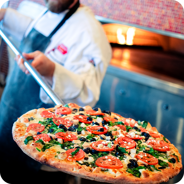 Pizza chef holding a baked pizza with tomatoes, olives, and greens near a hot oven.