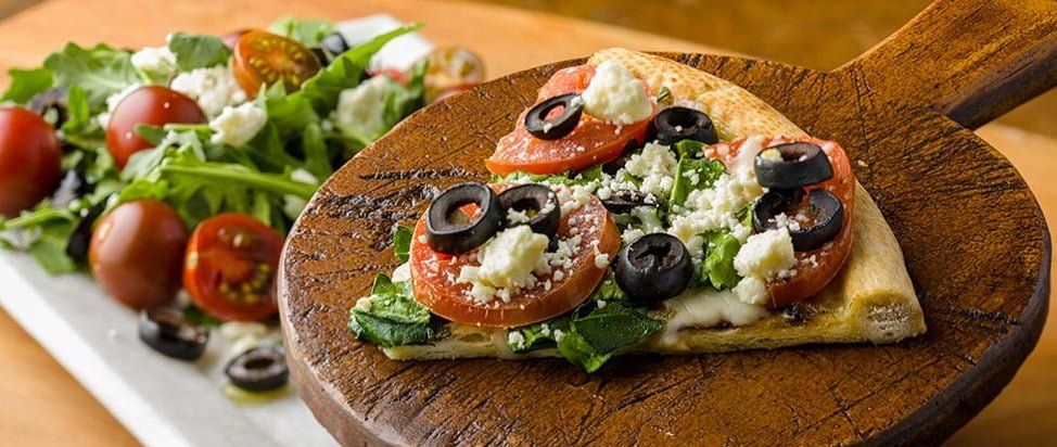 A slice of pizza topped with tomatoes, olives, and feta on a wooden board, served with a fresh salad.