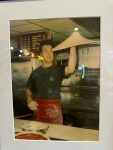 A man in a red apron tosses pizza dough. He stands in a commercial kitchen.