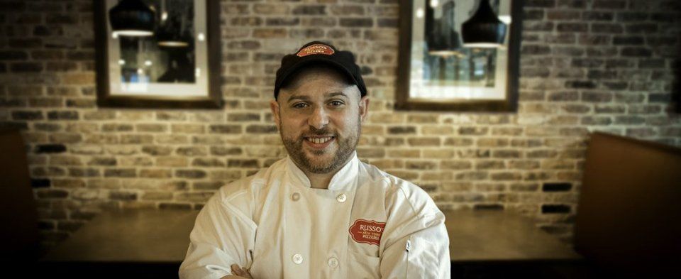 A chef in a white coat and baseball cap smiles at the viewer. He stands in a restaurant with brick walls and hanging lights.