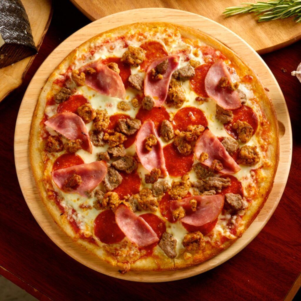 Pizza with pepperoni, ham, and sausage on a wooden board. The crust is golden brown.