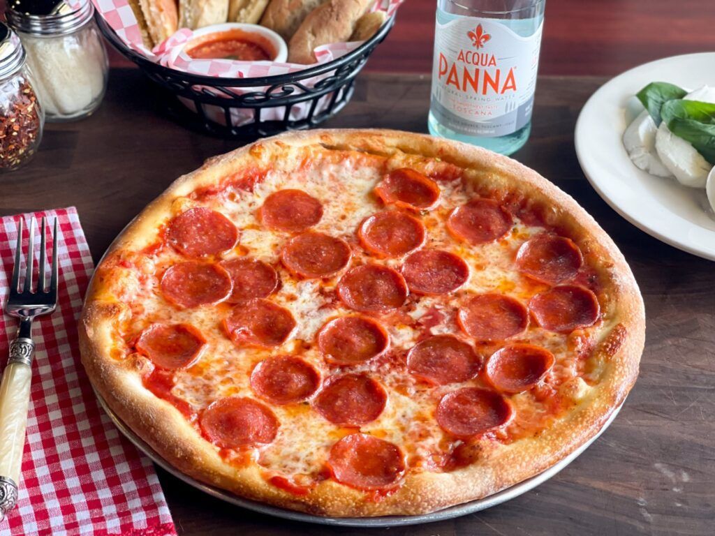 Pepperoni pizza on a metal platter, with breadsticks, water, and mozzarella on a wooden table.